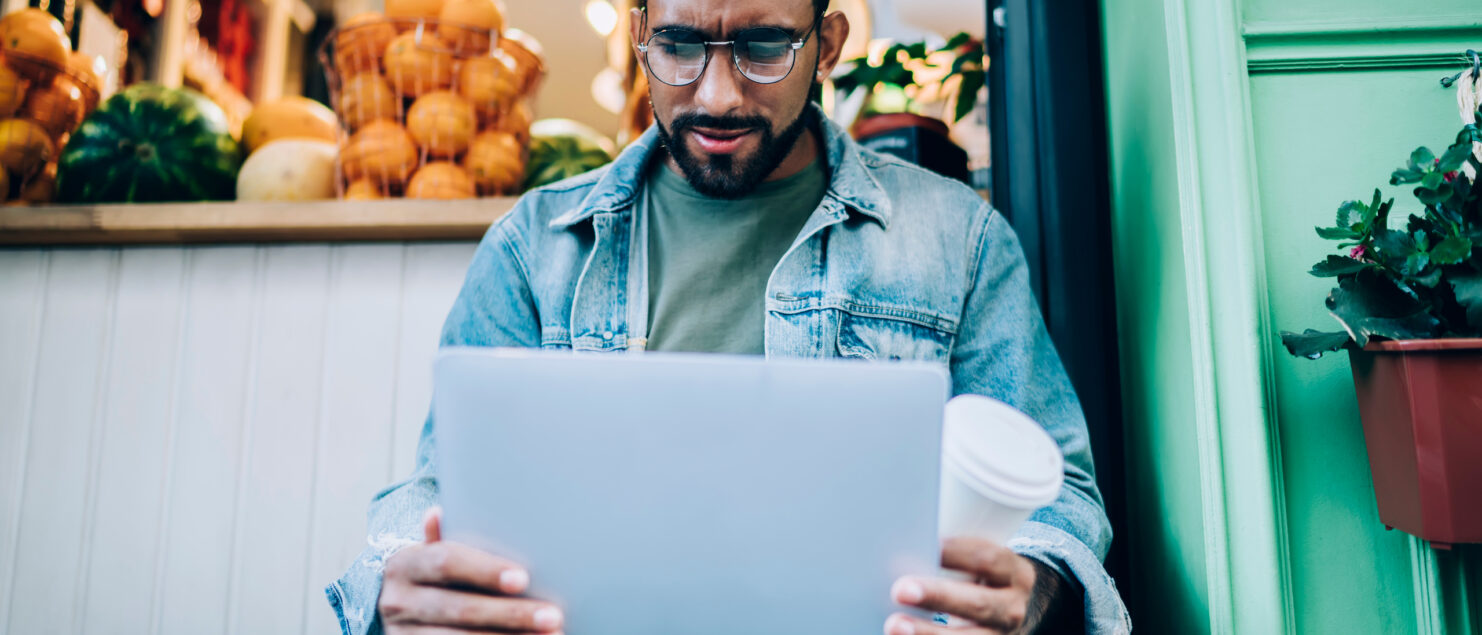 A man sits on a step looking at his laptop and holding a coffee cup in his hand.