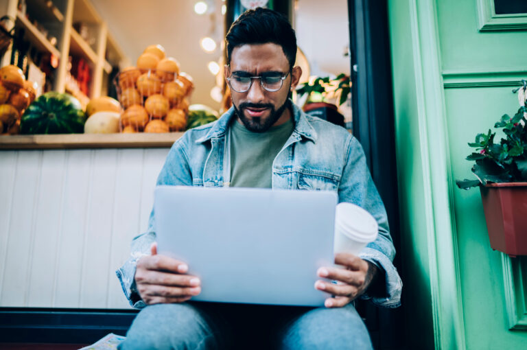 A man sits on a step looking at his laptop and holding a coffee cup in his hand.