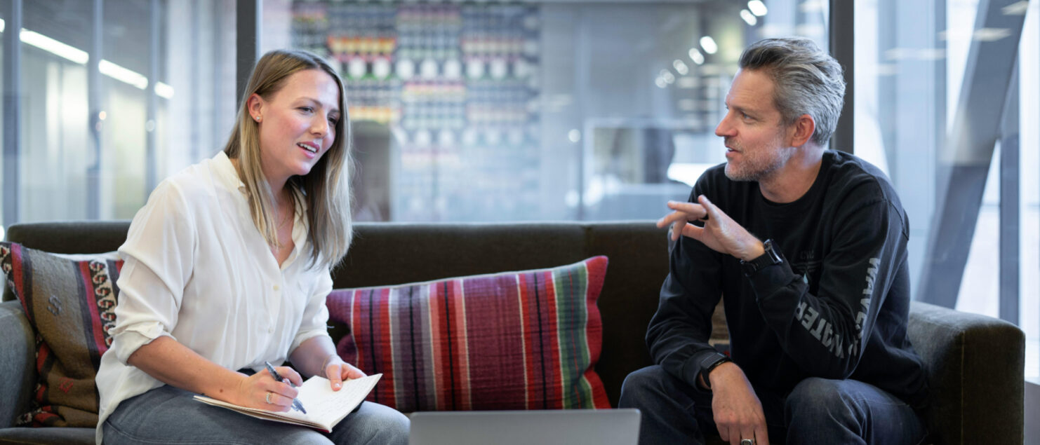 A professional man and woman sit on a sofa looking at a laptop, the woman makes notes on a notepad.