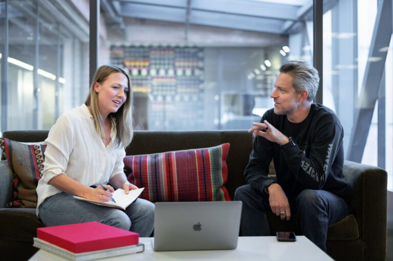 A professional man and woman sit on a sofa looking at a laptop, the woman makes notes on a notepad.
