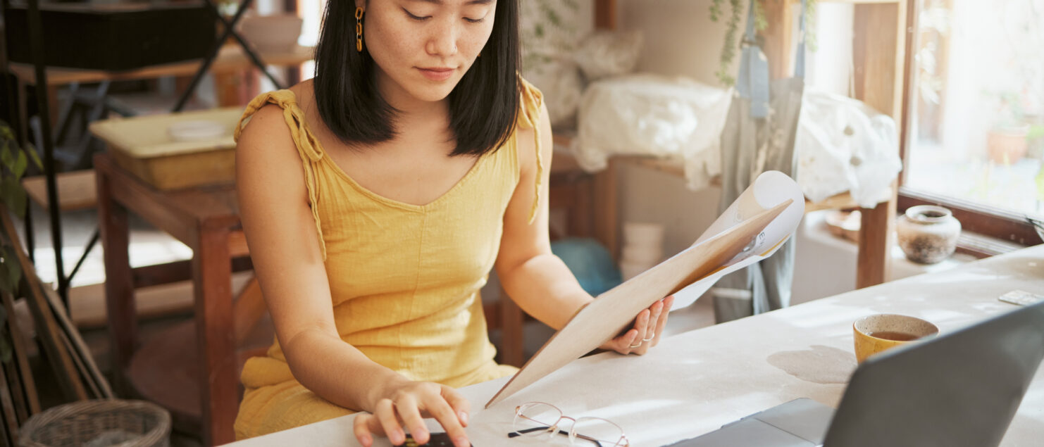 A woman sits at a desk in front of her laptop holding some papers and typing numbers into a calculator.