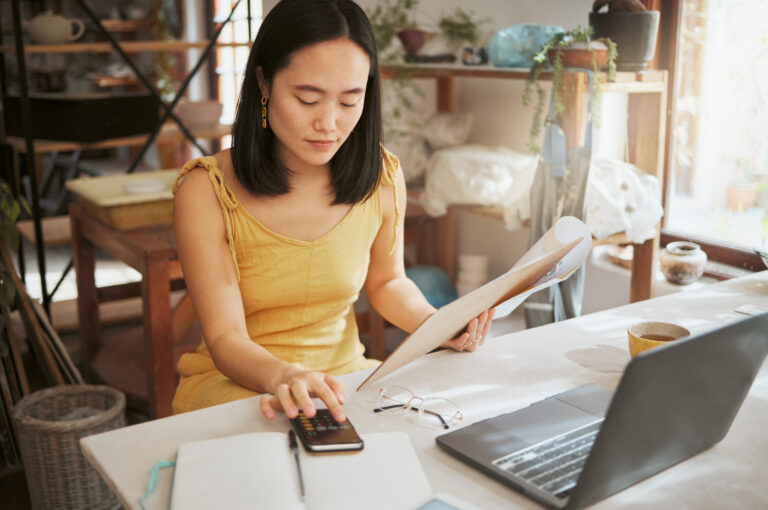 A woman sits at a desk in front of her laptop holding some papers and typing numbers into a calculator.
