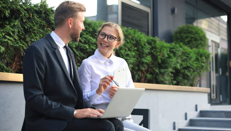 Two colleagues discuss their work outdoors with a laptop.